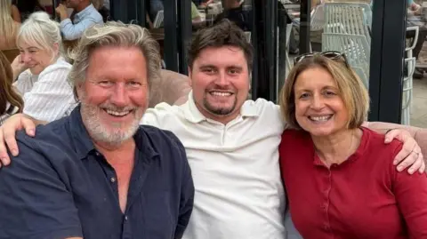 Alex Warwick A family group are all smiling to camera. Centre is a younger man with dark hair wearing a white polo shirt. He has his arms around the two on either side who are his parents. On the left is a middle aged man with grey hair and a beard, wearing a navy blue shirt. On the right is a middle aged woman wearing a red buttoned top. She has bobbed light brown hair and has sunglasses on her head. 