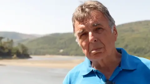BBC Man with blue polo shirt and greying temples looks towards camera, with sandbanks of an estuary behind him, and rolling green hillsides