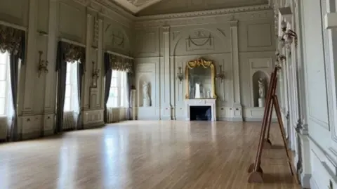 Essex Shire Hall Trust A regal and ornate looking hall which is empty. There are sage green walls, and curtain drapes above the windows.