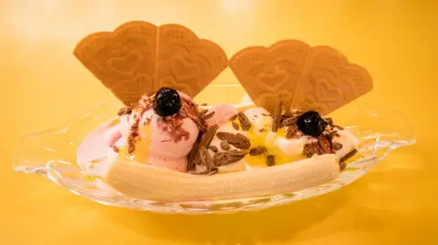 Emma Warley Warley's picture of a banana in a bowl alongside two scoops of ice cream. Two berries and wafers sit upright in the ice cream. Taken on a bright yellow table. 