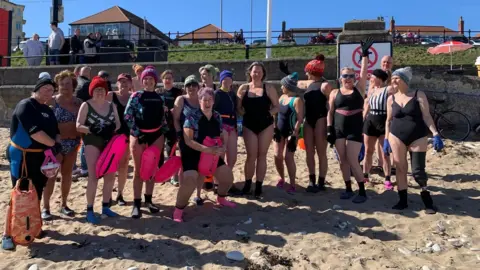 The Flamborough Flippers Sixteen ladies int their swimming costumes and winter hats are lined up on the beach for the photo. Brenda has gloves on and a black sleeve and sock on her leg.