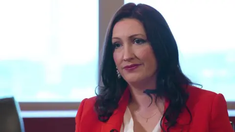 Emma Little-Pengelly is wearing a red blazer and white shirt. She has a fine necklace on and silver drop down earrings. She has long curly black hair and brown eyes. 