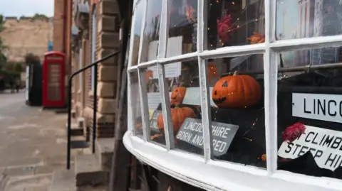 Visit Lincoln Side view of a shop window of three orange pumpkins with faces and signs such as 'Lincolnshire born and bred'. A telephone box can be seen in the distance and Lincoln Castle wall.