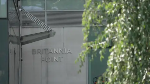 Exterior of Britannia Point in Colliers Wood, with the building's name visible on a grey wall behind glass-fronted facades and partially obscured by trees in the foreground.