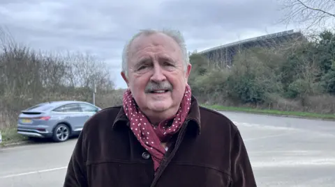 BBC/ Kate Bradbrook Man with grey hair, grey moustache who is wearing a red and white spotty scarf and brown jacket. He is standing near a road with a grandstand in the background.