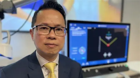 Supplied Dr Teik Choon See is wearing a blue blazer, white shirt and yellow tie. He has black hair and black glasses. He is looking at the camera while in front of a computer screen.
