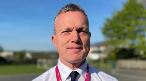 Nathan Cooper, who has greying short hair, is looking directly at the camera. He is smiling slightly and wearing a red lanyard around his neck, a white shirt and a dark tie. There is a very blue sky in the background and a blurred tree and playground.