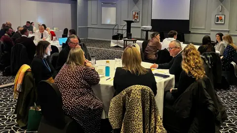 Lee Barron Smartly-dressed men and women sit at four round tables, with notepads, pens and water jugs on the table. They are in a hotel-style conference room with a highly-decorated black and white carpet.