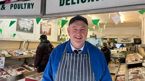 A middle aged man wearing a cap and royal blue butchers coat and blue and white striped apron smiles at the camera in front of his butchers shop in Brighton