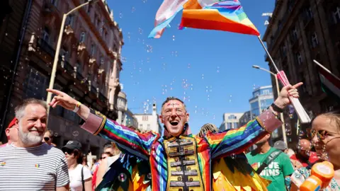 A man standing in the march wearing a multicoloured ringmaster type outfit. He has both his arms out and is holding a pride flag in his left hand. He is smiling and wearing glasses. To his left - in the bottom right corner of the picture - is a woman in sunglasses with a bubbles gun - there are bubbles across the scene. On his right, there is a smiling man in a white-and-black horizontally-striped t-shirt. He has two hear pride stickers on it.