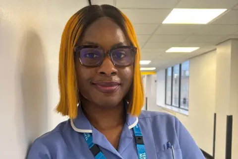 Nottingham University Hospitals NHS Trust A woman in a lilac nurses uniform. She is wearing glasses, has a light blue NHS landyard around her neck and is leaning against the wall of a hospital corridor.