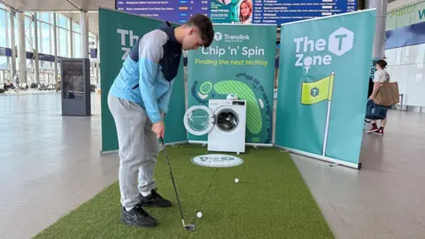 A white washing machine sits in the middle of a train station surrounded by green posters with 'chip n spin' written on it. A man is hitting a golf ball into the machine. He has dark short hair, grey jogging bottoms, black trainers and a blue, grey and navy jacket.