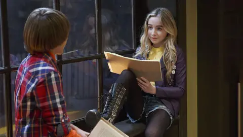 Getty Images Carpenter is sat on a window ledge holding a notebook sideways. She is peering over it at a young boy with long brown hair, who we can only see from behind as he faces Carpenter. He is wearing a red, white and blue checked shirt. Carpenter is wearing denim shorts with black tights and leather knee high boots with silver spikes. Her long blonde hair is down and in loose waves over her purple leather jacket, which has button badges on the lapel and is covering a yellow t-shirt.