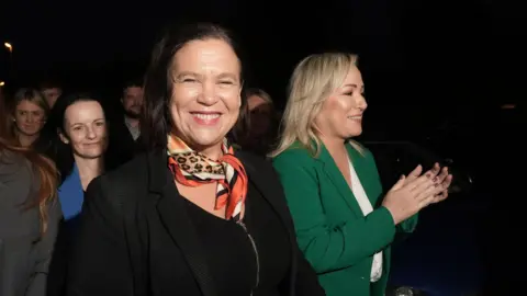 PA Media A woman with dark brown chin length hair smiles towards the camera. She is wearing a black top and jacket with a red patterned scarf. A woman with shoulder length blond hair stands to herright wearing a green blazer and white top. She is smiling to the right of the lens and clapping her hands.