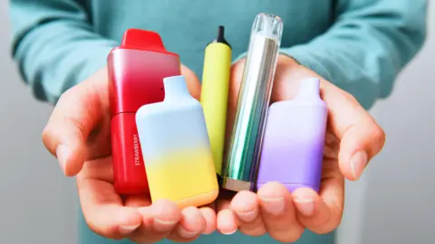 Getty Images A pair of hands is holding five disposable vapes of different colours, such as red, blue, yellow purple and multi-coloured. The person holding the vapes is wearing a turquoise jumper.