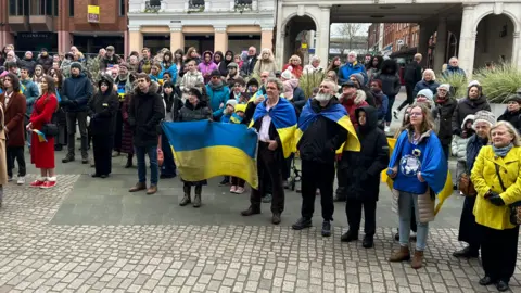 Richard Knights/BBC People standing in a town centre, many holding the Ukrainian flag. They appear to be listening to a speaker, who is out of shot. 