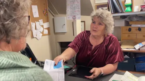 Shann Brede is standing behind the Post Office counter handing a document to a customer. Behind her there is a noticeboard, filing and a set of wooden drawers. To her right is a till and a jar of pens and there is paperwork to her left. The customer is obscured and only her grey hair and green jumper can be seen.