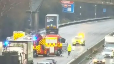 National Highways An overturned van can be seen just behind a fire truck. There are cars and lorries behind the emergency services. One person in a yellow high vis jacket is walking on the road.