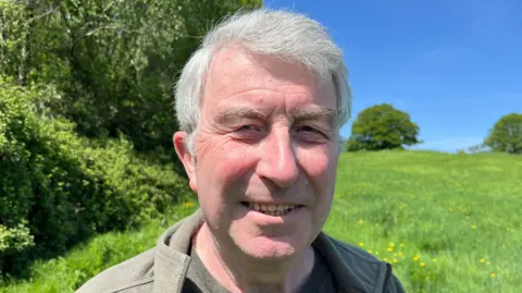 Steve has grey hair and is wearing a light green jacket. In the background are hedgerows and a green field with yellow flowers, with trees further away.