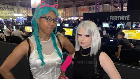 Two cosplayers stand side by side in a busy exhibition hall with rows of gaming monitors and a large Fortnite sign behind them. The woman on the left wears a turquoise braided wig with pink cat ears, glasses and a white corset top. The woman on the right wears a silver-white bob wig, red contact lenses, a black tactical outfit with a gold chain and pink gloves.
