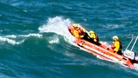 RNLI/Lizzie W An orange inshore RNLI boat is seen on the crest of a wave with three volunteers seen on board.
