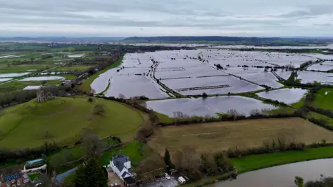 An areal shot of fields covered by water. Glastonbury Tor, a ruin at the top of a hill, is visible to the left of the image.