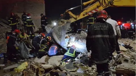 Emergency personnel wearing uniforms and hard hats search in rubble with a digger at night time. 