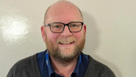Derrick Roberts is standing against a shiny white school wall. He is bald with a ginger beard and is wearing glasses. He is also wearing a dark grey jumper and a shirt with a lanyard around his neck