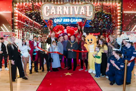 Carnival Golf and Games A crowd stands on a red carpet outside the entrance of Carnival Golf and Games. A man in mayoral chains cuts a red ribbon with others holding baloons and cheering.