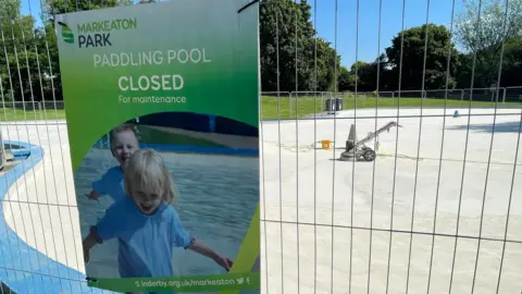 Fencing and an information sign around a closed paddling pool with repair equipment in the background