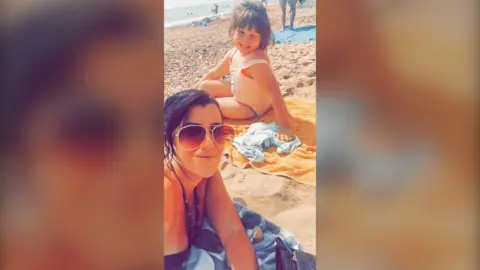 Family Handout/PA Real Life Rebecca takes a selfie with Summer, they are sitting on towels on the beach, both looking at the camera. Summer is further away from the camera, she's sitting on an orange towel, wears a pale pink swimming costume and is smiling at the camera, she has dark blonde hair tied up with a fringe. Rebecca is closer to the camera, she is on a blue patterned towel and wears a strapped black swimsuit and large aviator style framed sunglasses, she has black hair which is tied back and a side fringe.