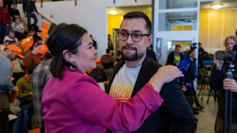 Getty Images MUTE EGEDE fica em uma sala movimentada cheia de pessoas e equipes de câmeras como um político - uma mulher vestida de rosa - rostos em sua direção, com a mão no ombro dele 