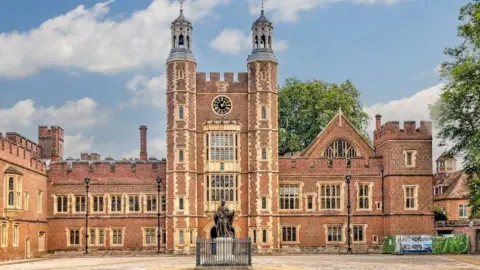 Getty Images An Eton College building constructed of red brick with a clock tower in the middle and a black statue on a white plinth in front of the entrance.