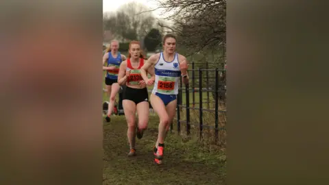 Ian Dunn Three women running on the course. The one in the foreground is wearing a white and blue Sedgefield Harriers vest with blue running briefs on and an orange race bib. Behind her is a woman with red hair in an orange and black running kit on. There is a woman behind her who is in blue and yellow with blonde hair.