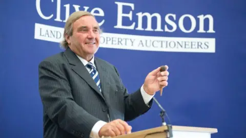 Clive Emson holding a gavel while standing at a lectern. He is wearing a dark thin-striped suit and a striped blue and white tie.