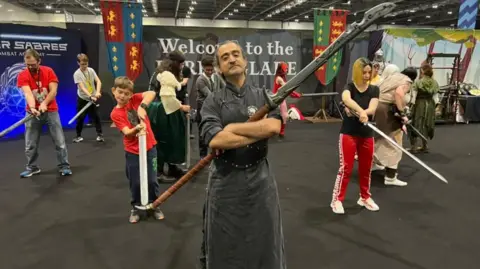 Thomas Copeland / BBC News Several people wield swords behind a man in the centre, who has his arms crossed over a larger sword.