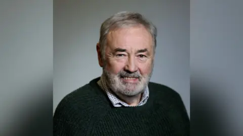 John Allen. He has white hair and beard and brown eyes. He is wearing a green jumper with check shirt underneath.