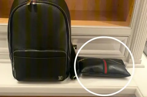 Black Louis Vuitton backpack with gold zippers on a white shelf, next to a small Gucci black pouch with green and red stripes and gold zippers, highlighted by a white circle.