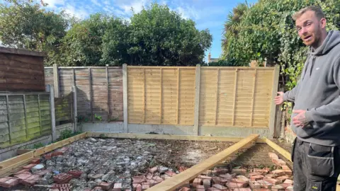 Gemma Hemmings Steve Figg, who is wearing a grey hoodie and trousers, pointing at the foundations of what was to become the garden office.