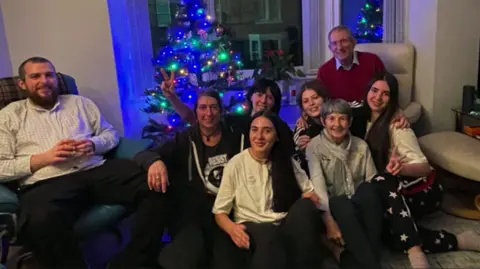 A group of eight sits on the floor in front of a Christmas tree. They look at the camera and smile.