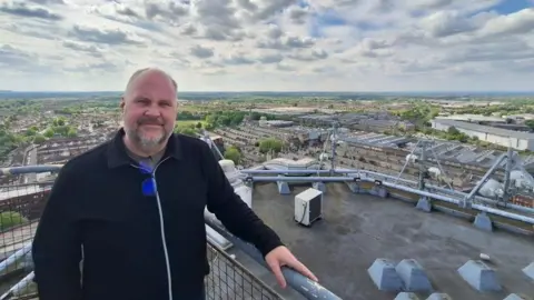 Jim Robbins smiles at the camera wearing a black jacket. Behind him, a wide bird's-eye view of Swinond, a variety of buildings, trees and parks with the countryside in the background.