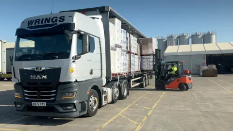 A lorry in a yard with a fork-lift truck loading boxes on to the back.