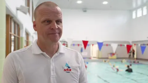 A man with close-cropped fair hair, who is wearing a white polo shirt with "STA" logo, stands in front of a swimming pool in a large white-walled building. Red, white and blue flags hang across the width of the pool and people can be seen swimming in the background, which is out of focus.