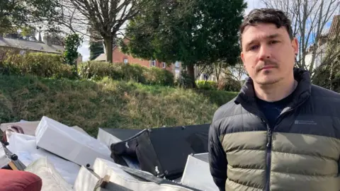 A man with dark hair in a green and black coat in front of a pile of rubbish in the street