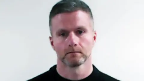 A police mugshot of a man with short grey hair in a black top