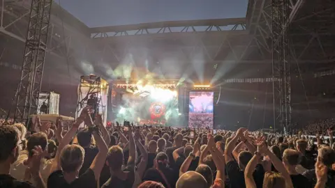 Paul K A large crowd gathered in front of a stage where Guns N' Roses are performing. People have their arms up in the air. It is night time and the strobe lights are lighting up the arena.