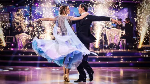 BBC Karen Carney and Carlos Gu dancing with their arms wide, smiling at each other. Carney wears a clitter light blue dress while Gu is wearing a black suit with his black hair slicked back.
