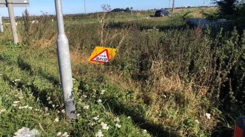 JOHN DEVINE/BBC A new give way sign lying among grass and other plants in a dip/verge at Boots Bridge crossroads