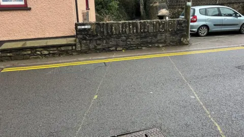 A road in front of a small stone wall with yellow lines drawn to mark where the culvert is 