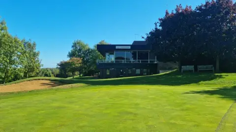 Tony Ashforth A golf clubhouse with large windows and an elevated terrace overlooking the course. Mature trees frame the scene on both sides, while a sand bunker is visible to the left of the building. Bright sunshine and a clear blue sky illuminate the landscape, highlighting the neatly cut grass and surrounding greenery.